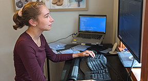 A woman works on a desktop computer in her own office with a laptop beside her.